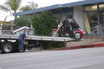 2 men are seen holding a motorcycle upright on a flatbed truck while a third works the controls of the bed lift.