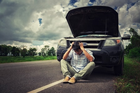 A man sits with his hands on his head in front of his broken down car with the hood open on the side of the road.