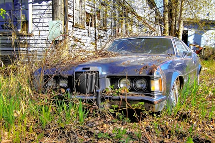 A blue junk car sitting with grass and weeds covering it, in front of an old house.