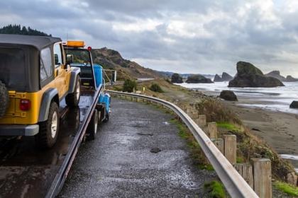 An orange jeep being loaded onto a flatbed truck overlooking a shore.