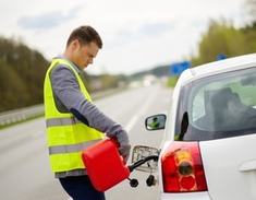 A tow truck operator is refilling the gas of a white car on the side of the road with a small gas can.